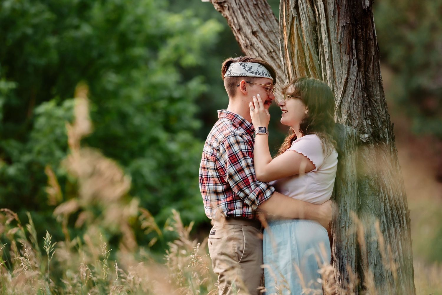 A romantic couple shares tender moments in a natural outdoor setting with trees and tall grass in summer.