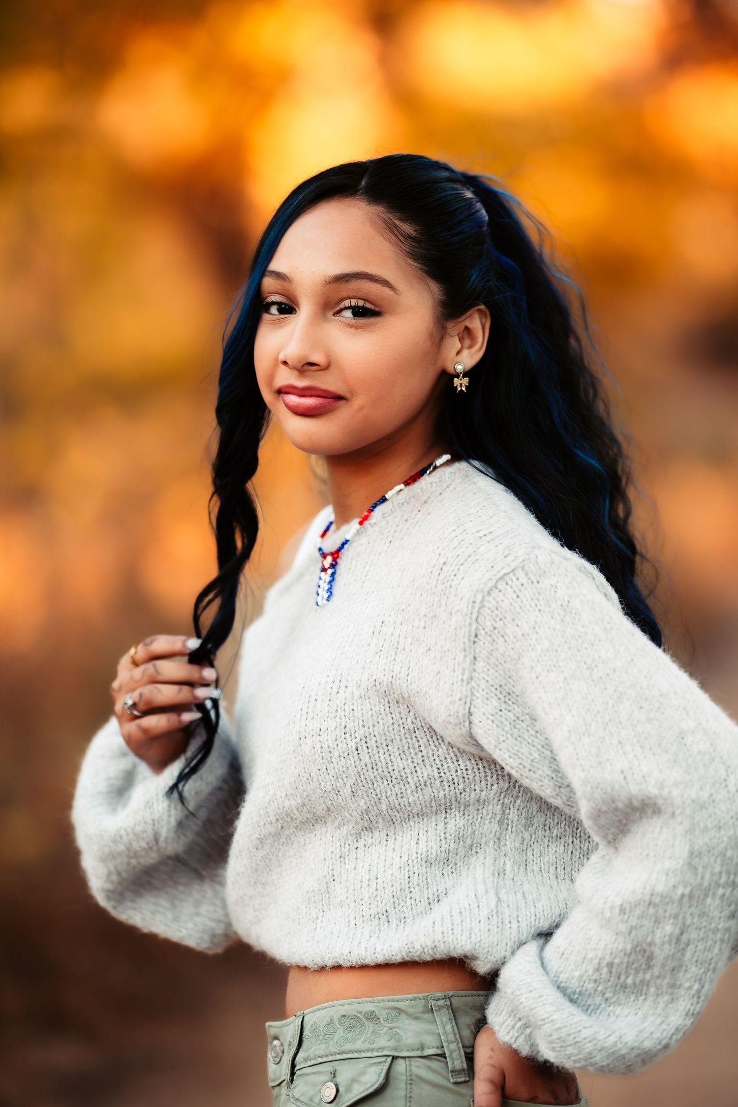 Woman in cozy white sweater and olive pants poses against warm autumn sunset background.