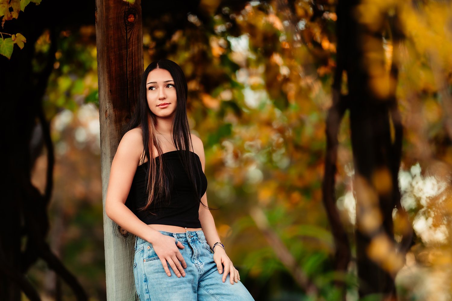 A person in a black top and jeans poses outdoors against autumn foliage with warm golden bokeh lighting.