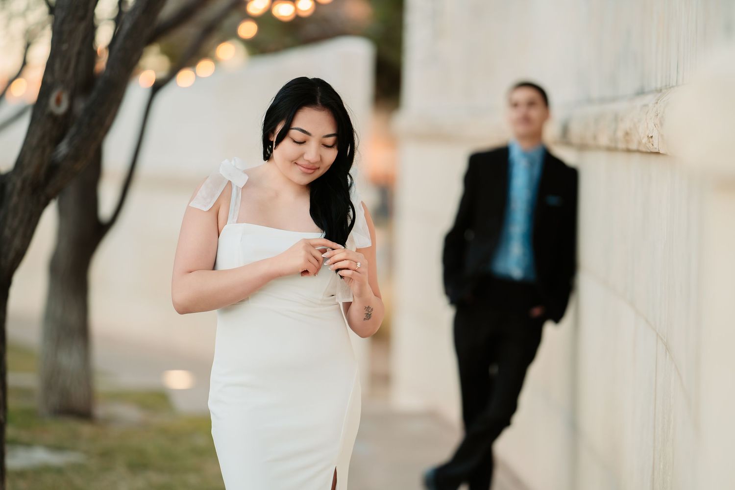 Elegant couple in formal attire pose outdoors with string lights and trees in background at dusk.