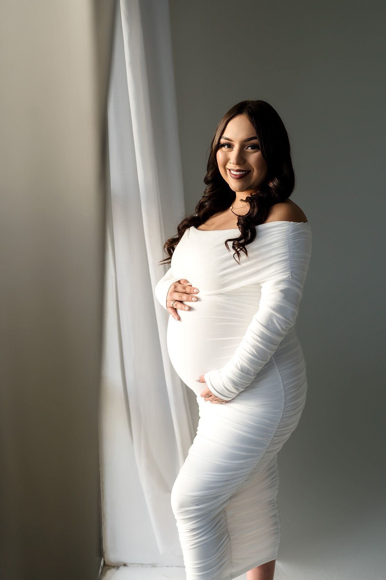 Smiling pregnant woman in white ruched off-shoulder dress posing by sheer curtains in a bright studio setting.
