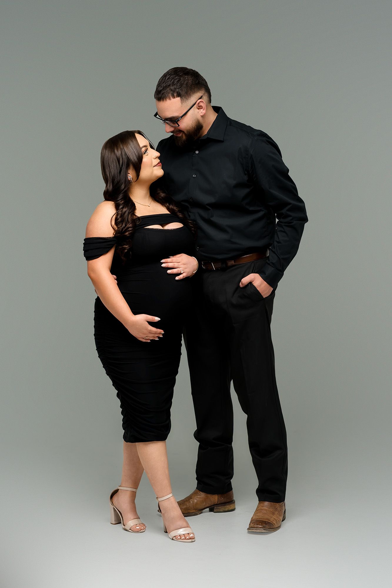 Pregnant couple in matching black outfits posing together on gray background, man leaning in toward woman.