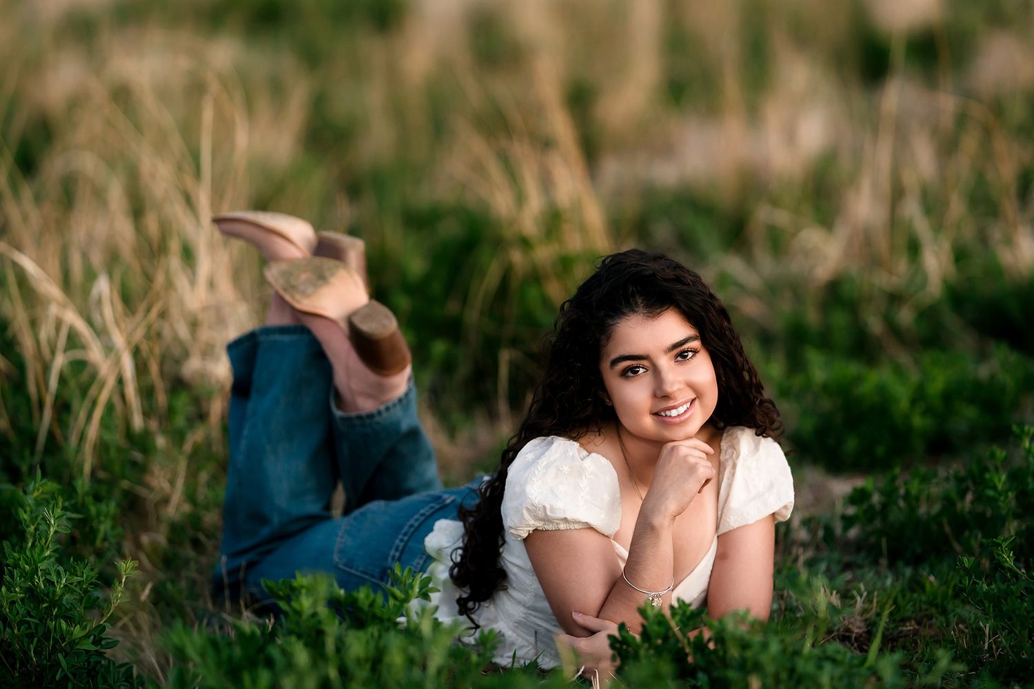 Young woman in white top and jeans lying in green grass field, smiling warmly at camera during golden hour.