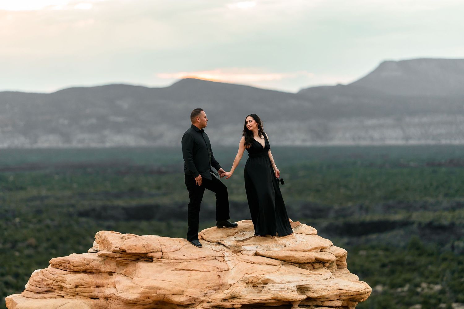 Couple in black attire holding hands on a red rock cliff with mountain views at sunset.
