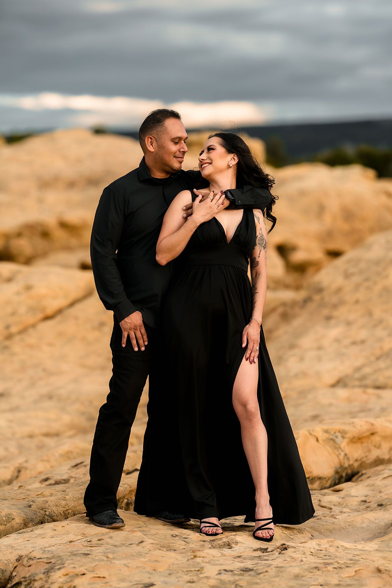 Elegant couple in black attire smiling together on rocky terrain with stormy sky backdrop.