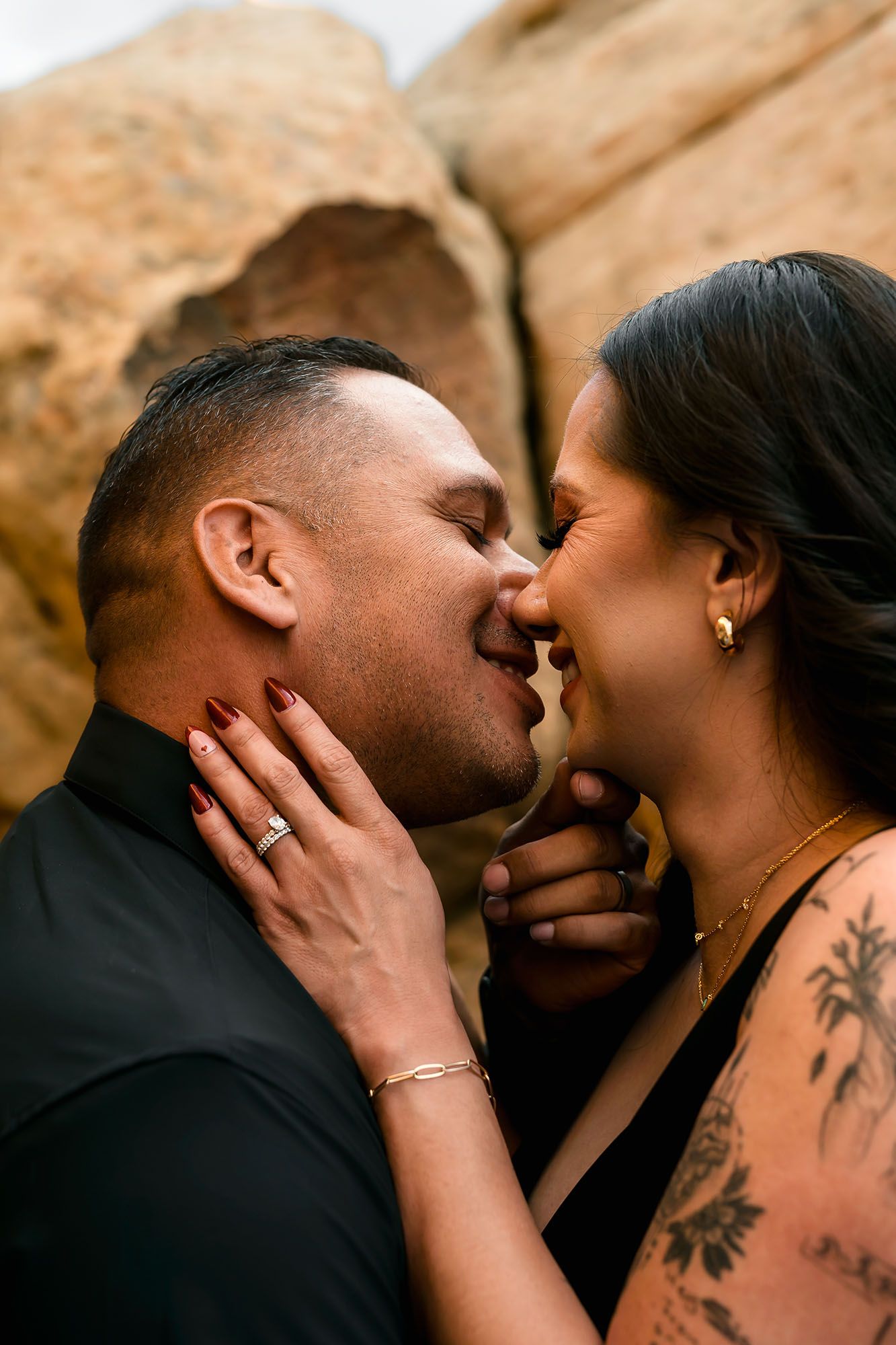 Romantic couple kissing with eyes closed, woman's tattooed arm embracing man in black shirt outdoors.
