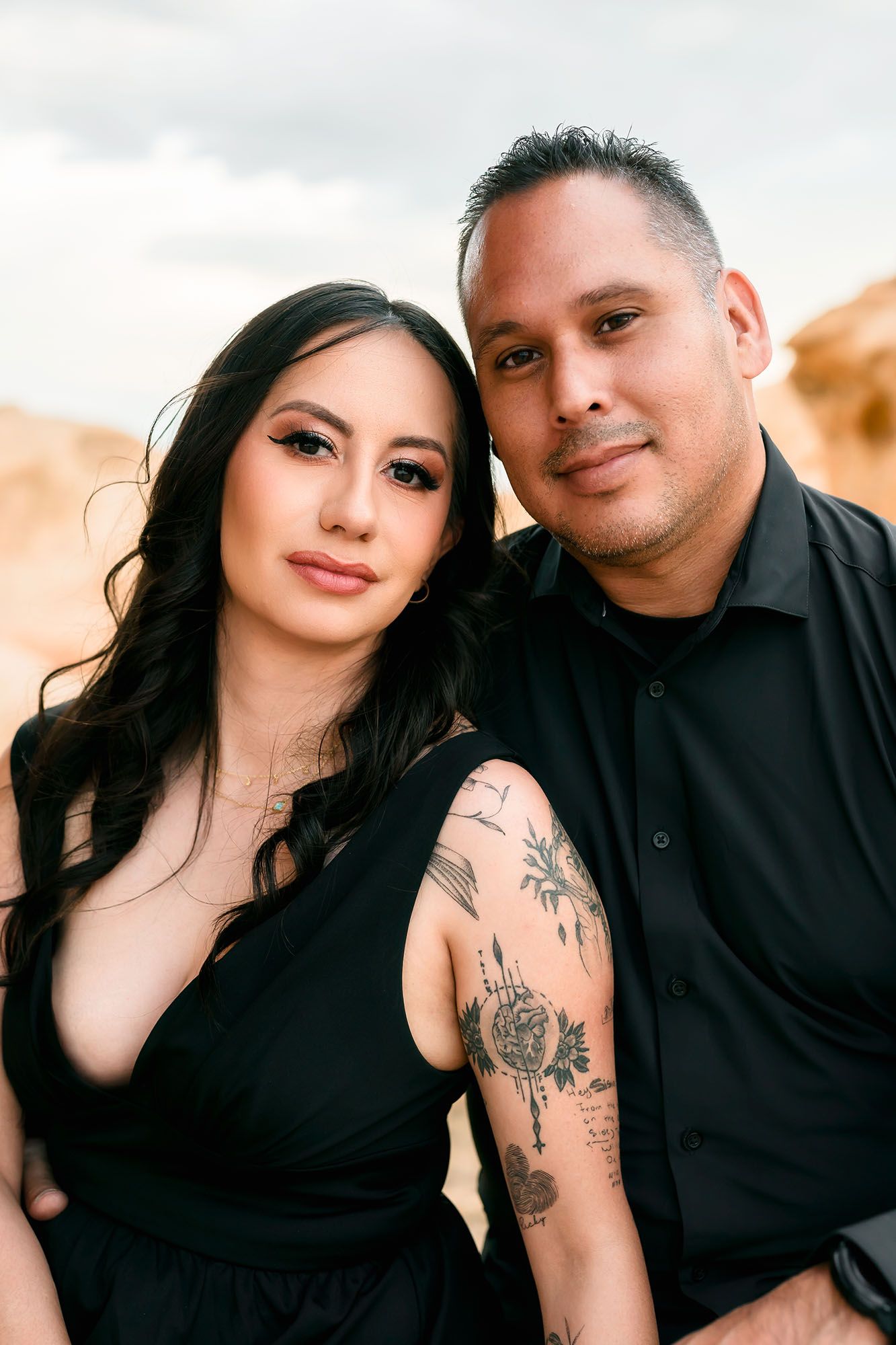 Close-up portrait of couple in black outfits, woman with floral arm tattoos leans on mans shoulder, outdoor setting.