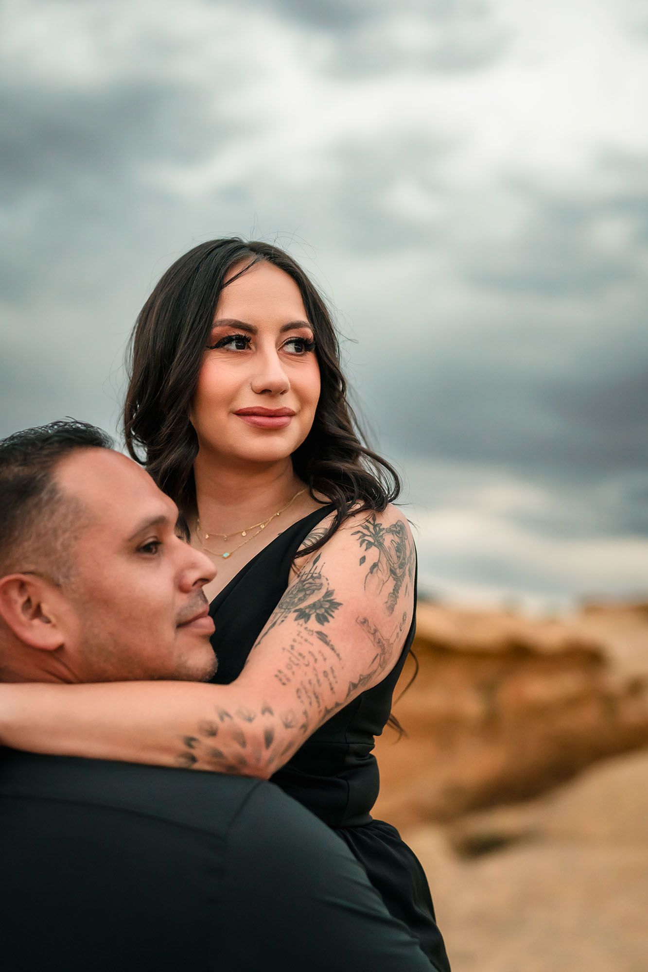 Woman with floral arm tattoos embracing man on desert rocks under dramatic storm clouds.