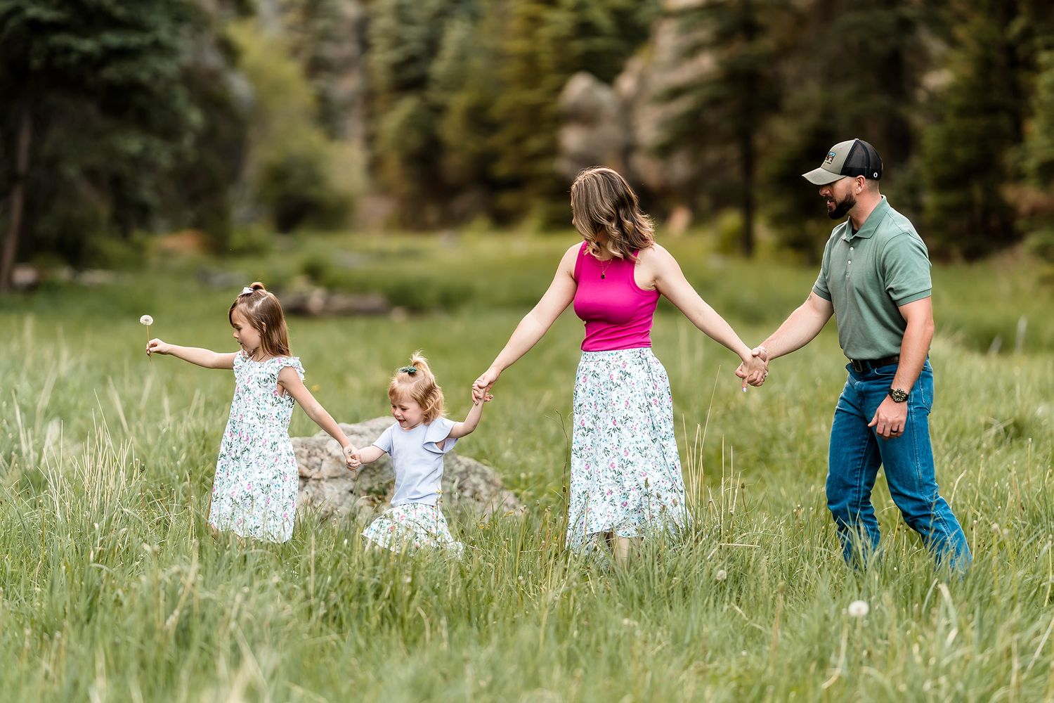 Family of four holding hands in a green meadow surrounded by pine trees, two young girls in floral dresses with parents.