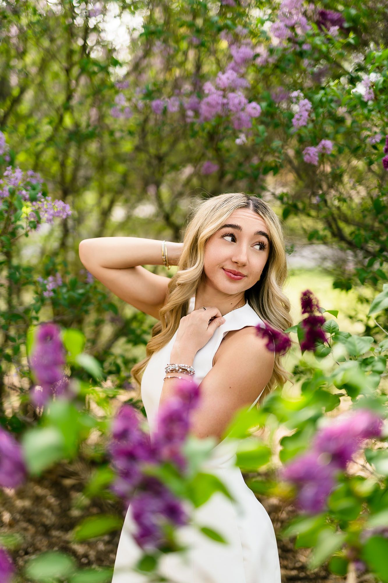 Blonde teenage girl in white dress posing among purple lilac flowers in green garden.