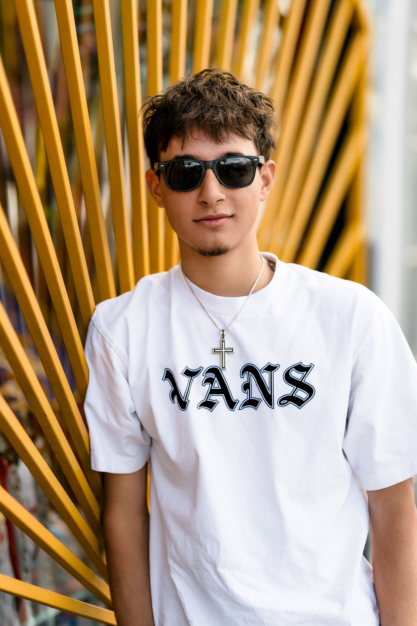 Young man in white Vans t-shirt and sunglasses poses with cross necklace against yellow geometric background.