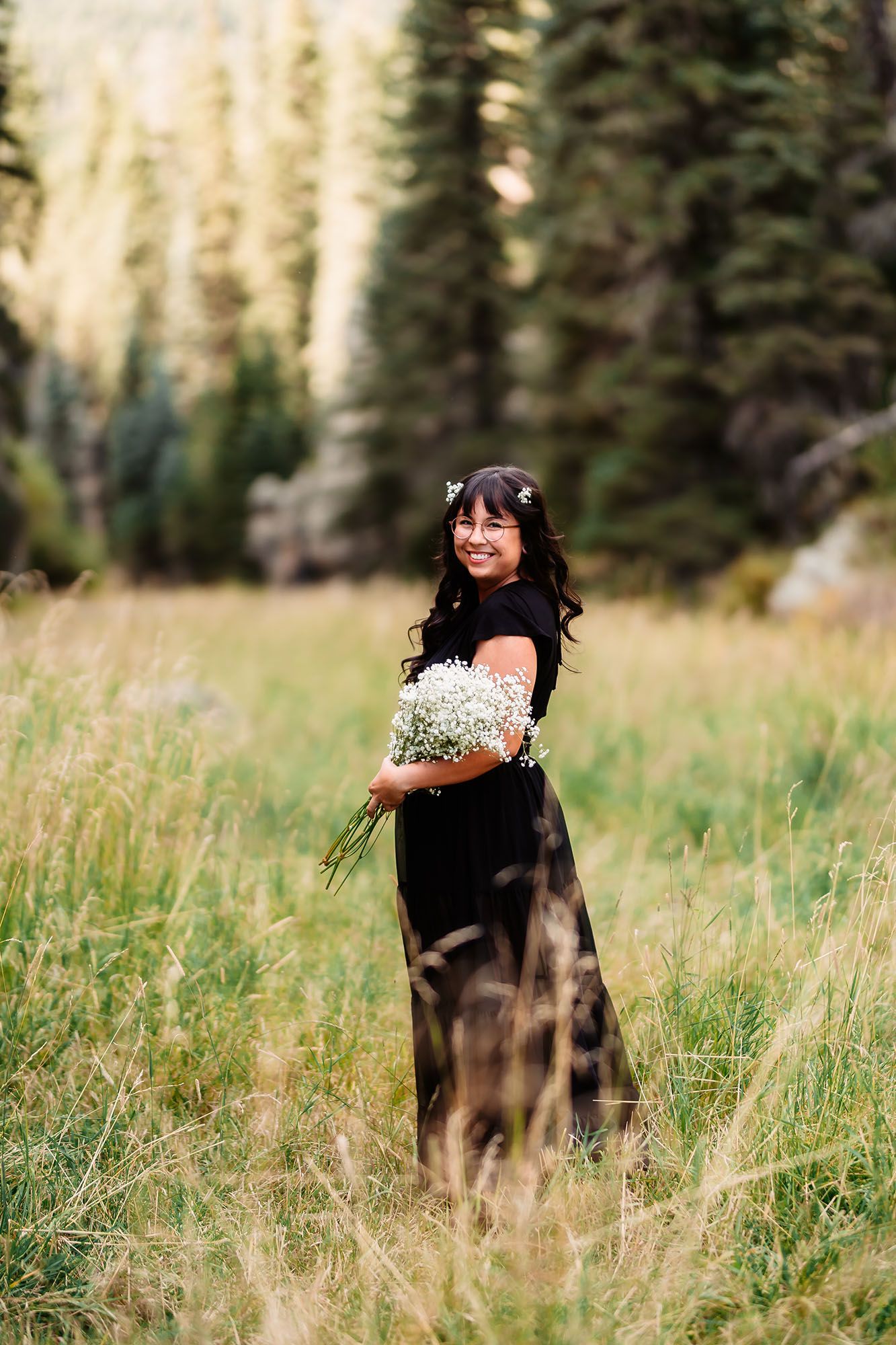 Woman in black dress holding white flowers stands in a meadow with pine trees in background.
