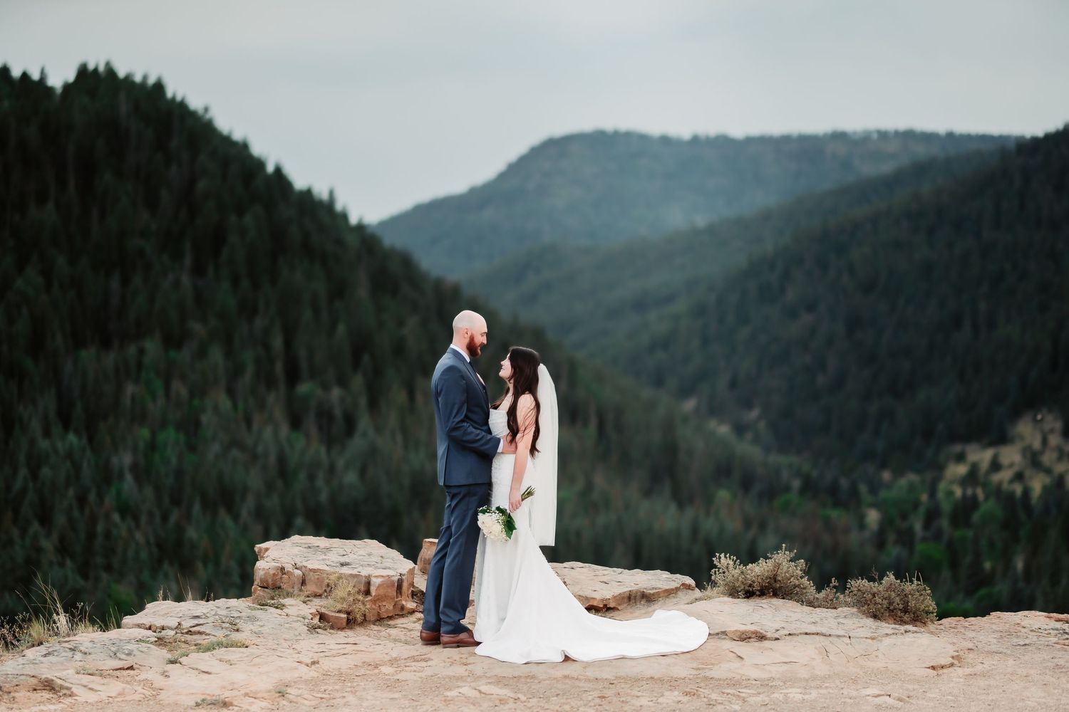 A couple in wedding attire photographed on top of a mountain overlook with forest-covered peaks in the background.