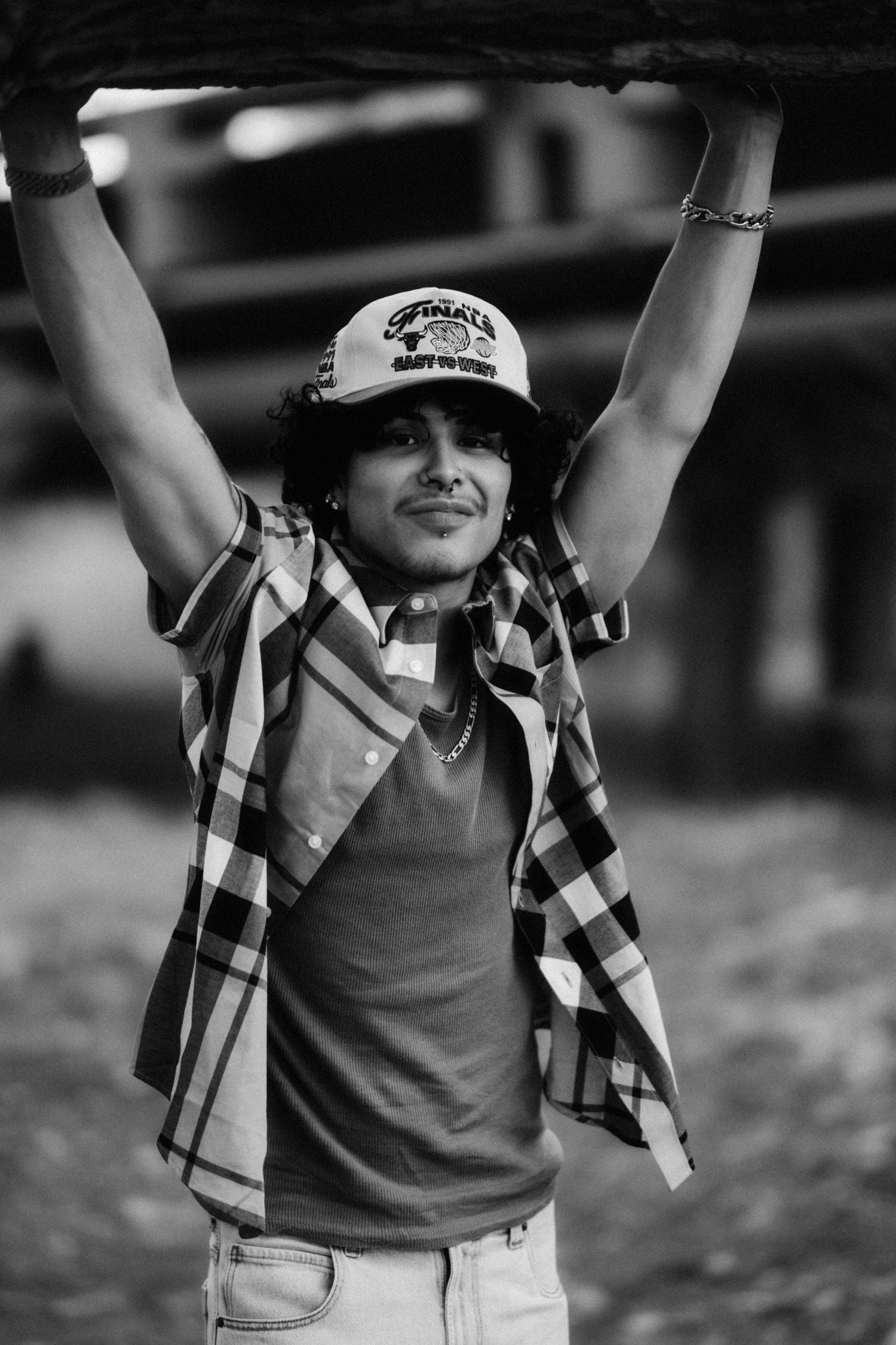 Black and white portrait of someone wearing a baseball cap and plaid shirt with arms raised joyfully outdoors.