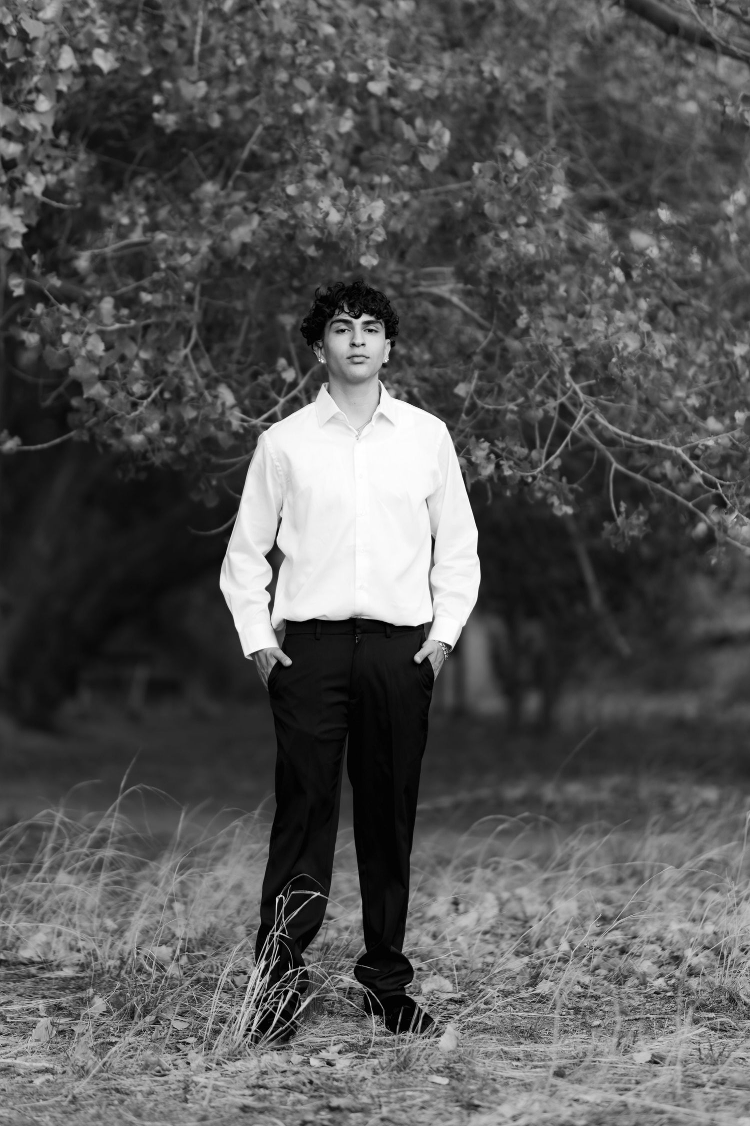 Black and white portrait of someone in white shirt and dark pants standing in field with trees in background.