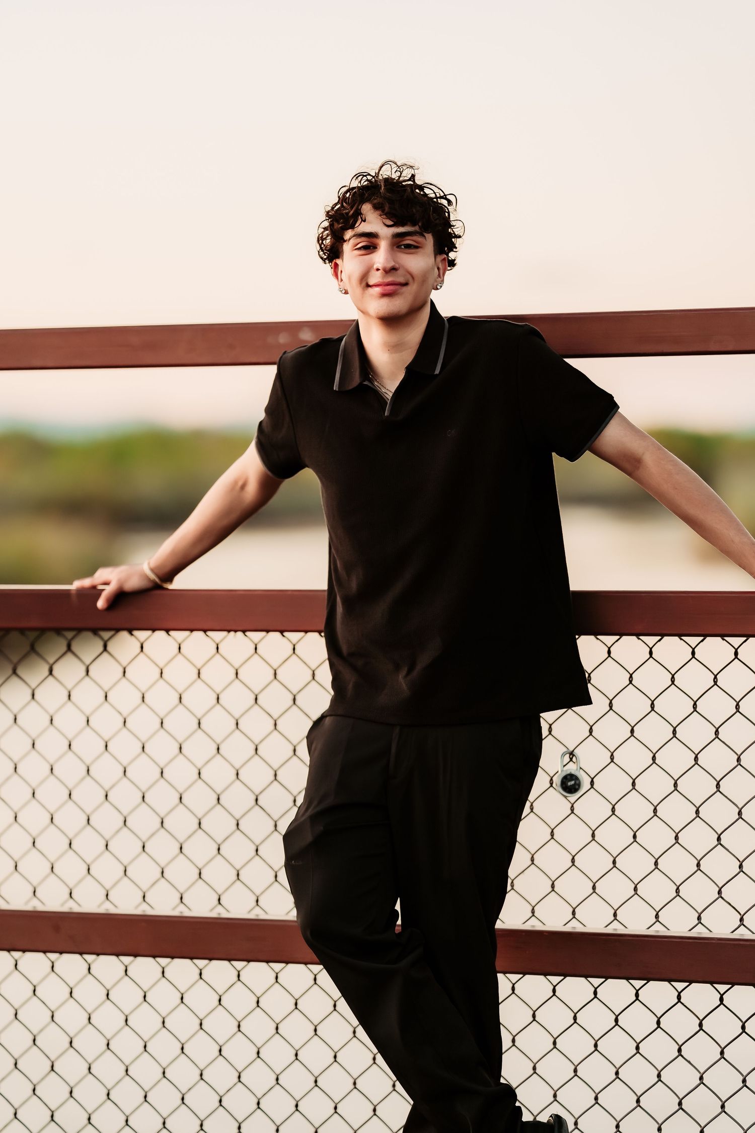 Person in black polo leaning against chain link fence at sunset with relaxed pose.