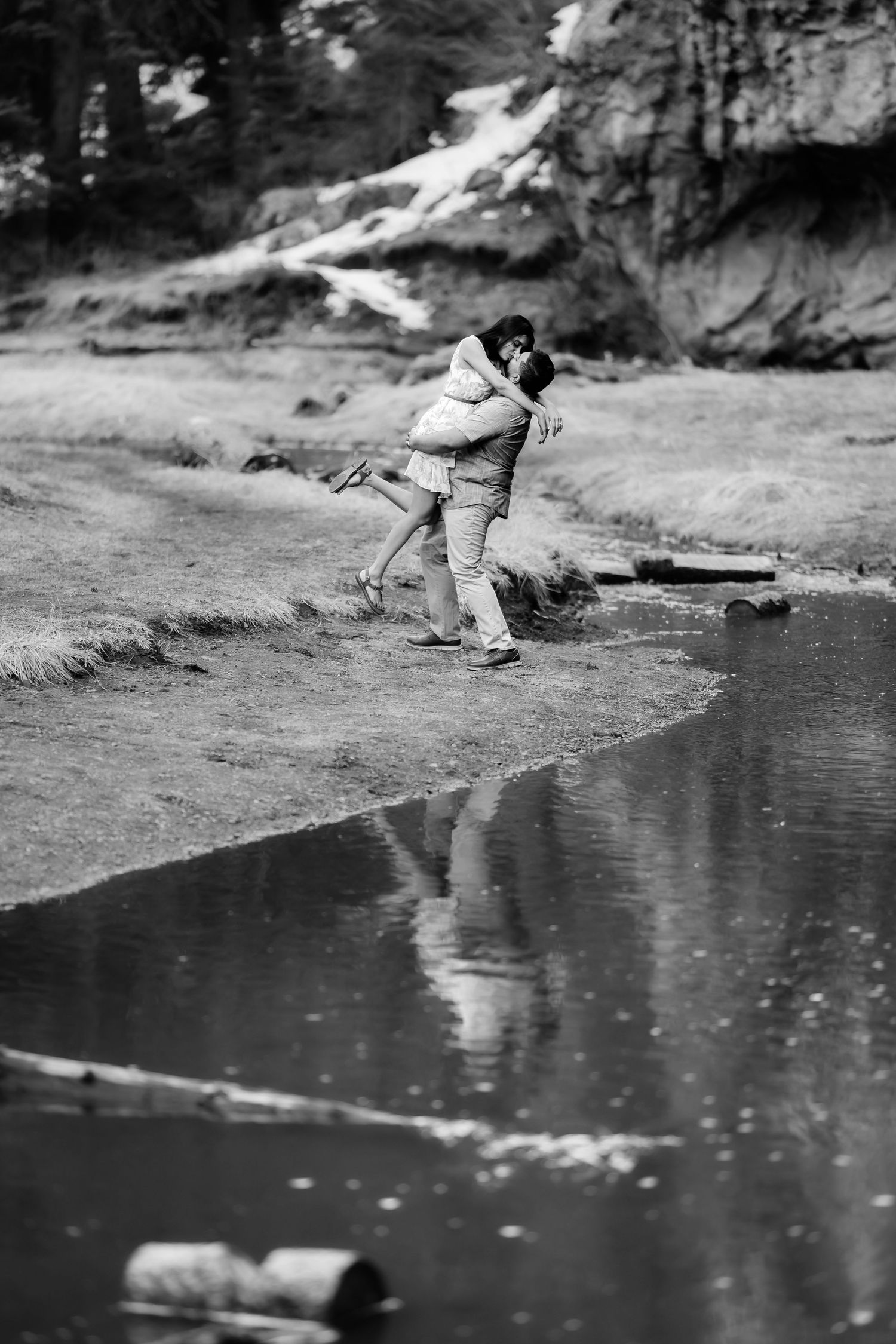 A black and white photo captures a romantic moment by a reflective stream with cliffs in the background.
