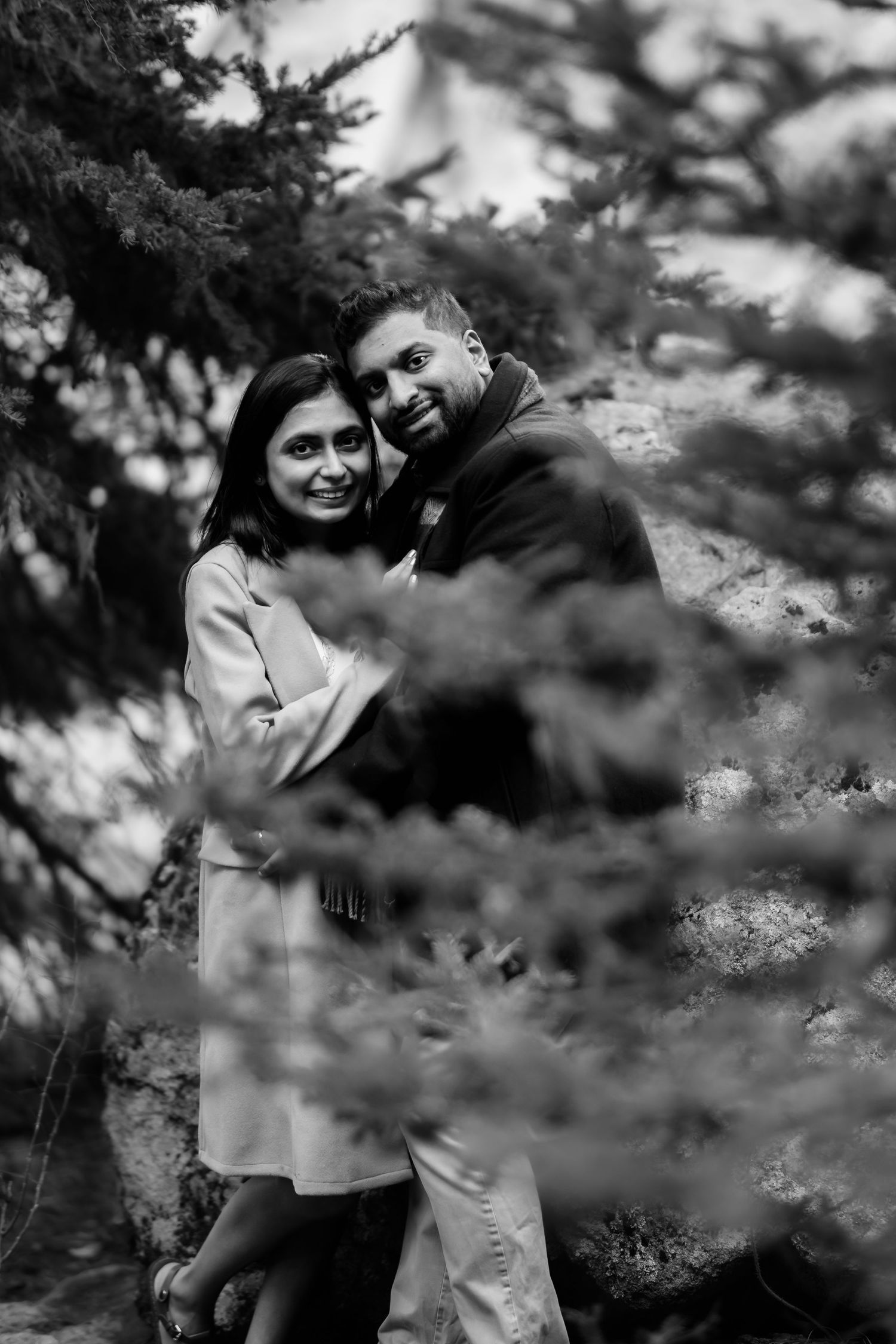 Romantic black and white portrait of a couple embracing amongst evergreen branches in a moody outdoor setting.