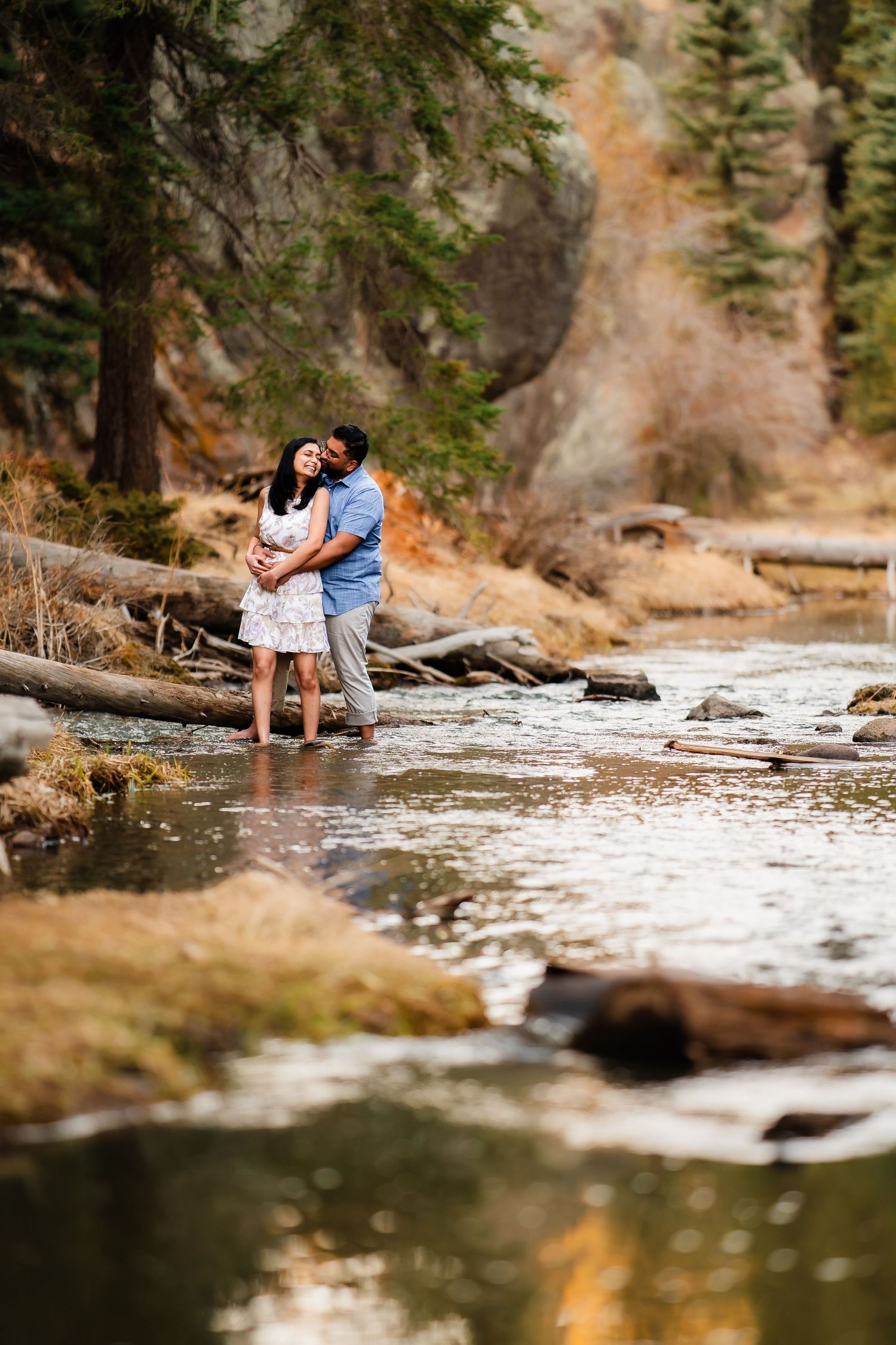 A couple shares a romantic moment while standing at the edge of a river surrounded by forest and rocky cliffs.