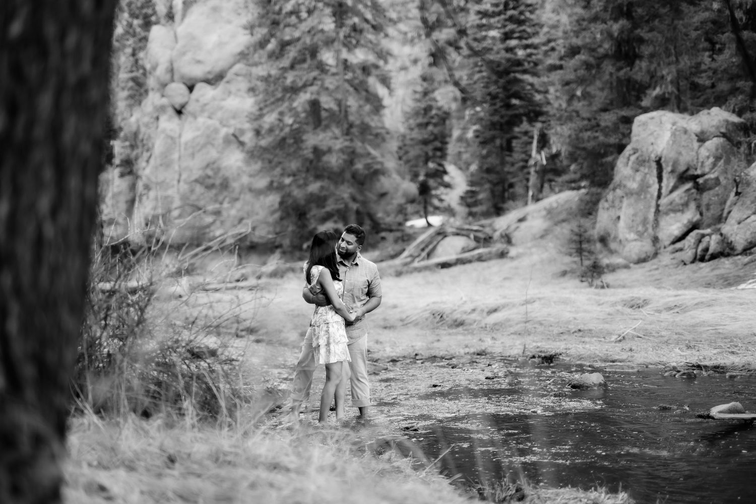 A black and white image of a couple embracing by a serene lake surrounded by trees.