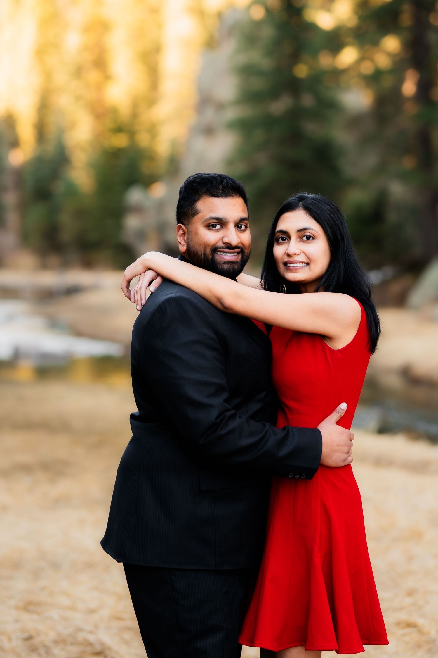 A couple in coordinated red and black formal wear embrace while posing for a sunset engagement photo.