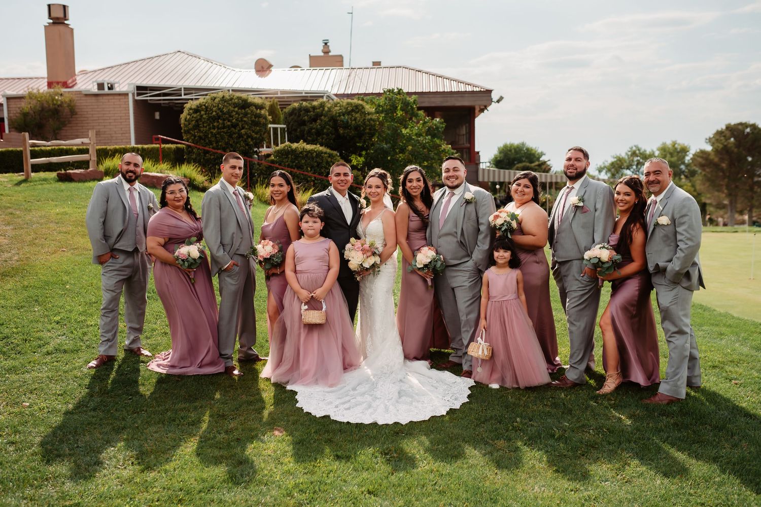 A wedding party poses together on a lush green lawn wearing dusty rose and gray attire.