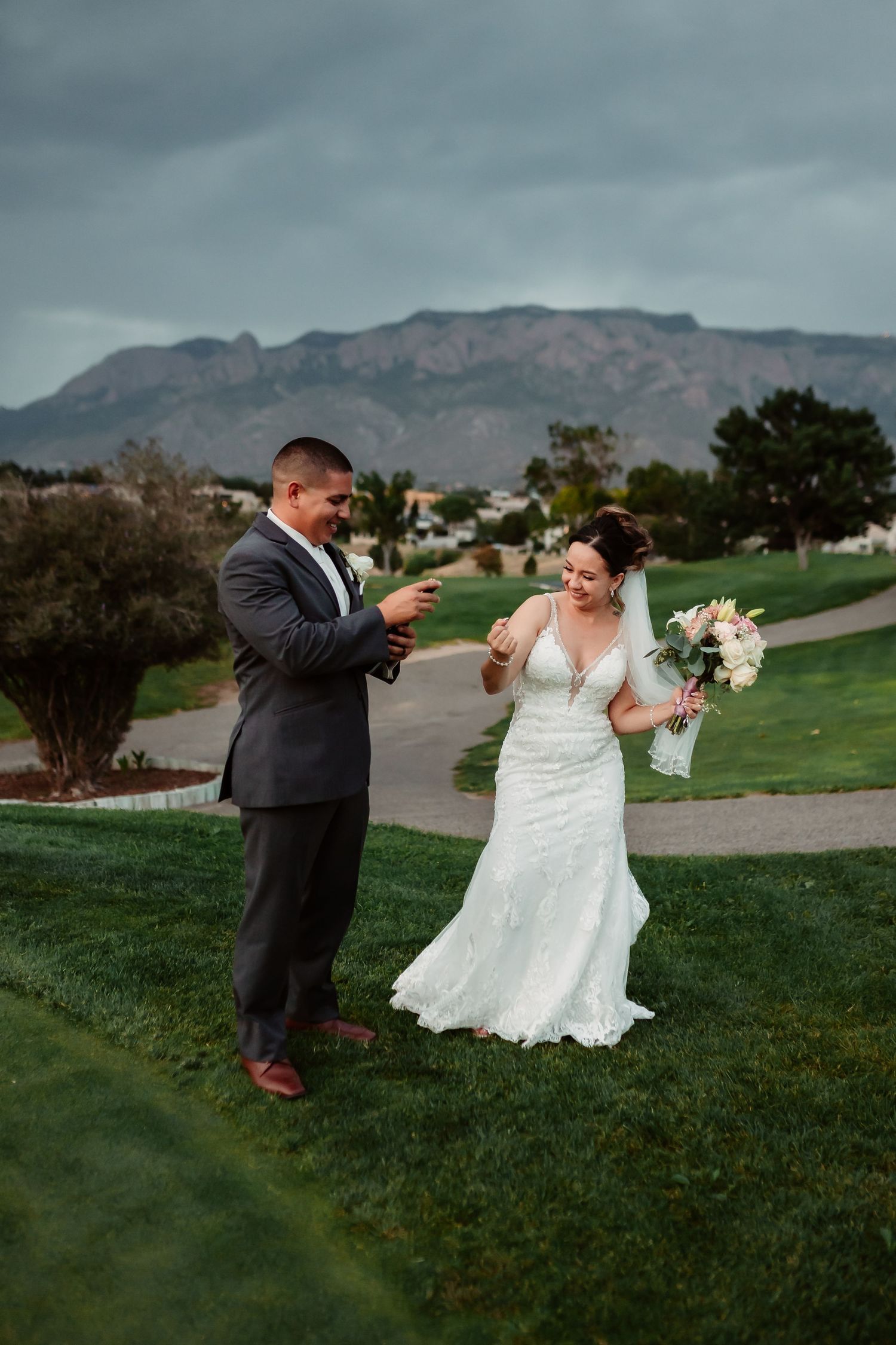 A playful wedding couple dances and poses on a grassy hill with mountains in the distance.