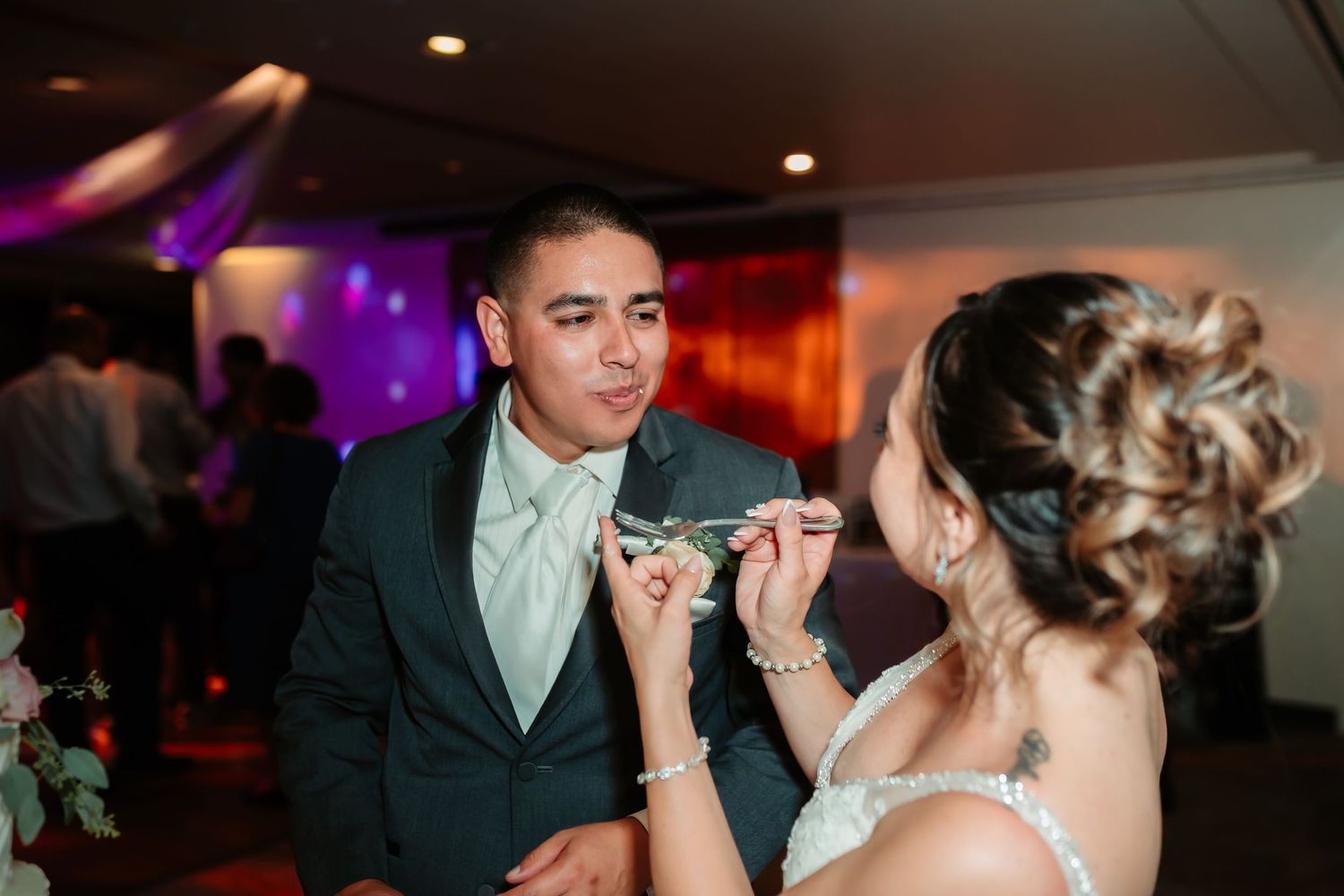 Couple sharing wedding cake at reception with purple ambient lighting in background.