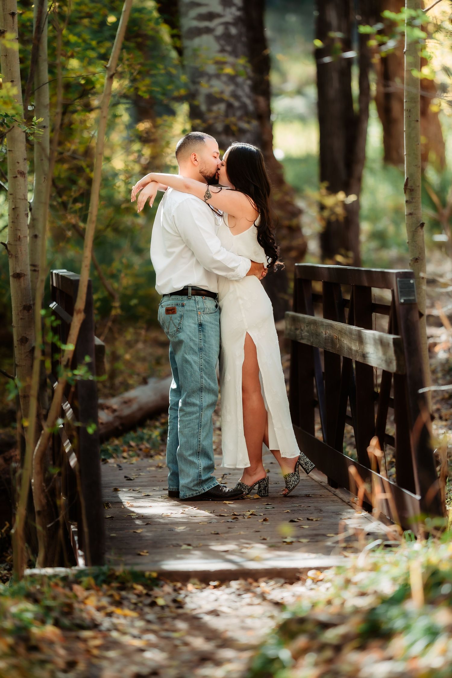 A couple shares an intimate moment on a rustic wooden bridge surrounded by lush forest greenery in natural lighting.