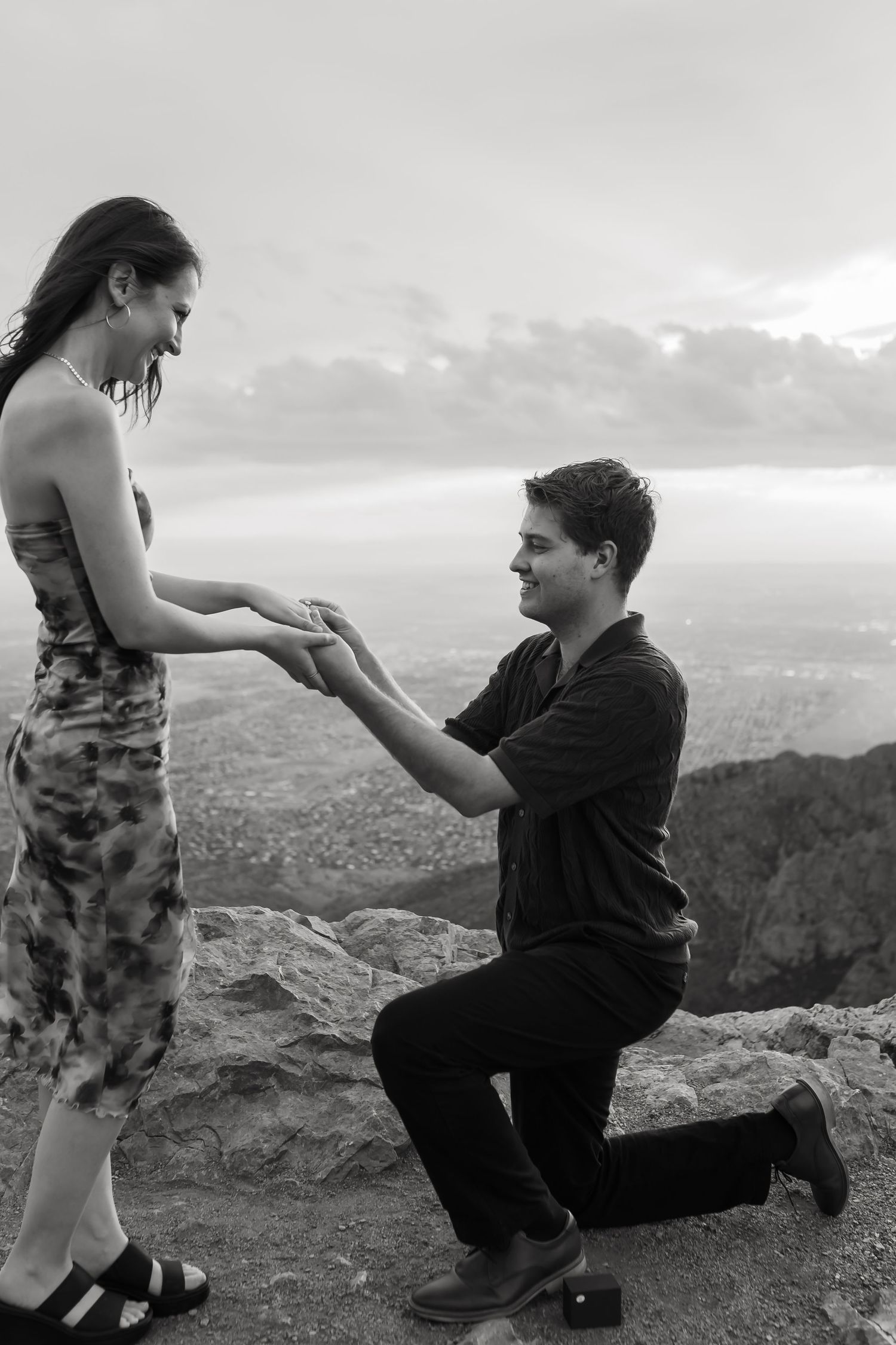 A couple shares a romantic proposal moment on a scenic mountain overlook at sunset.
