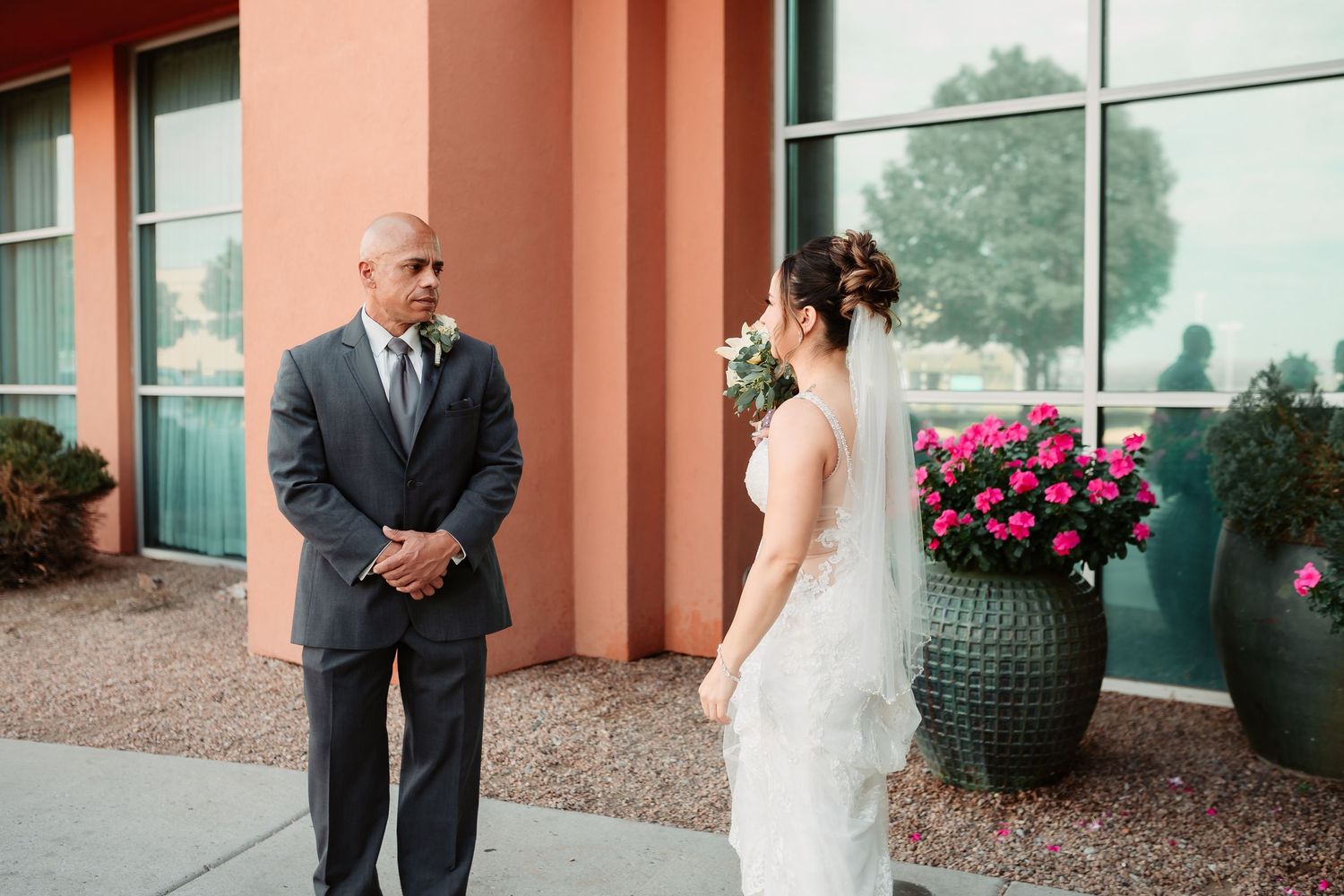 Emotional first look between wedding couple in front of coral-colored building with pink flowers and glass windows.