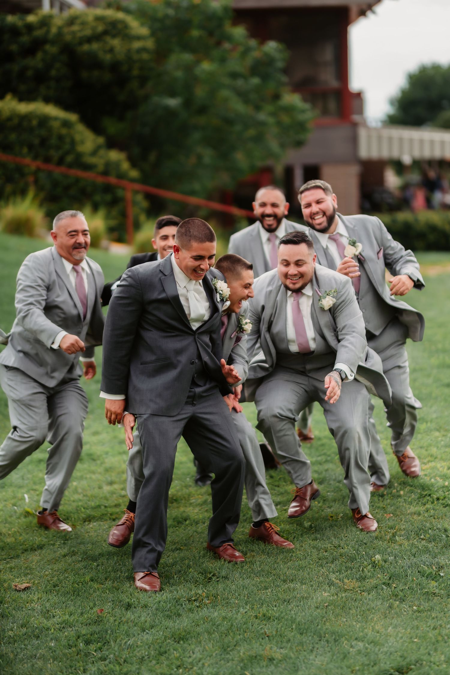 Wedding party members in matching gray suits perform a synchronized crouch pose on grass.