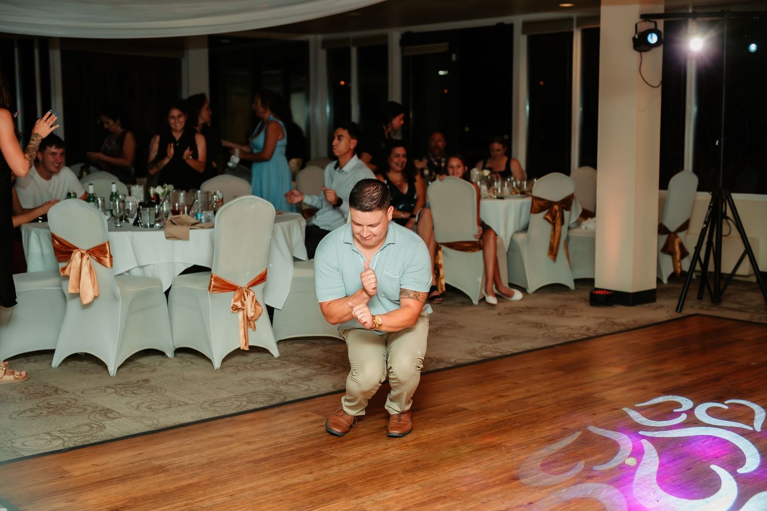A wedding guest performs a dance move on the hardwood floor while other guests watch from decorated tables.