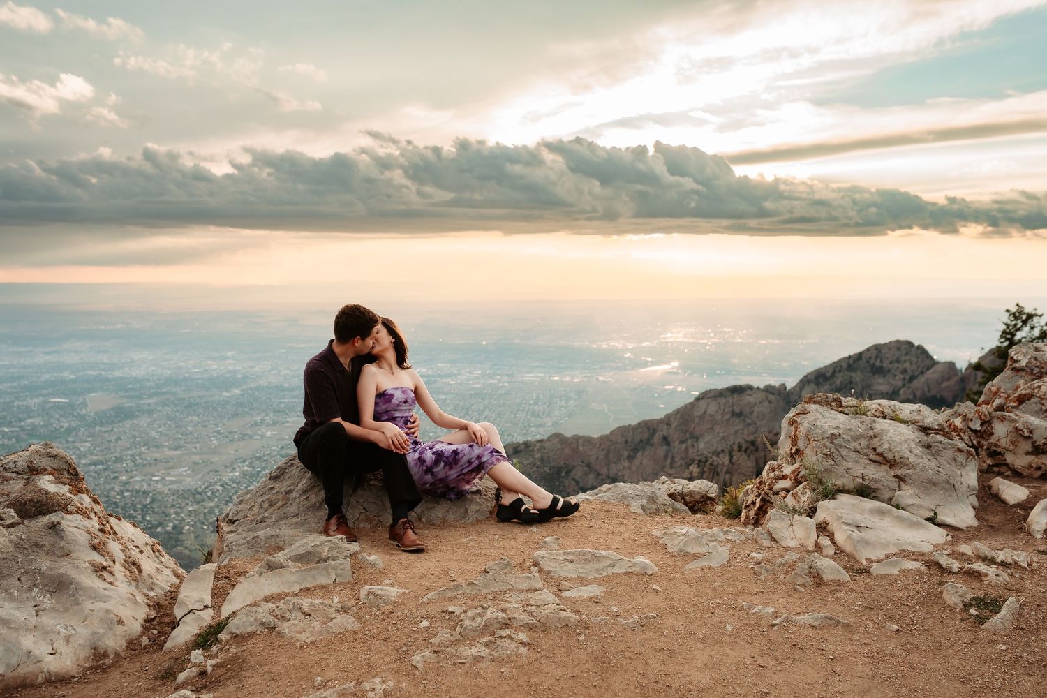 Couple embraces on scenic mountain overlook with expansive valley views at golden hour.