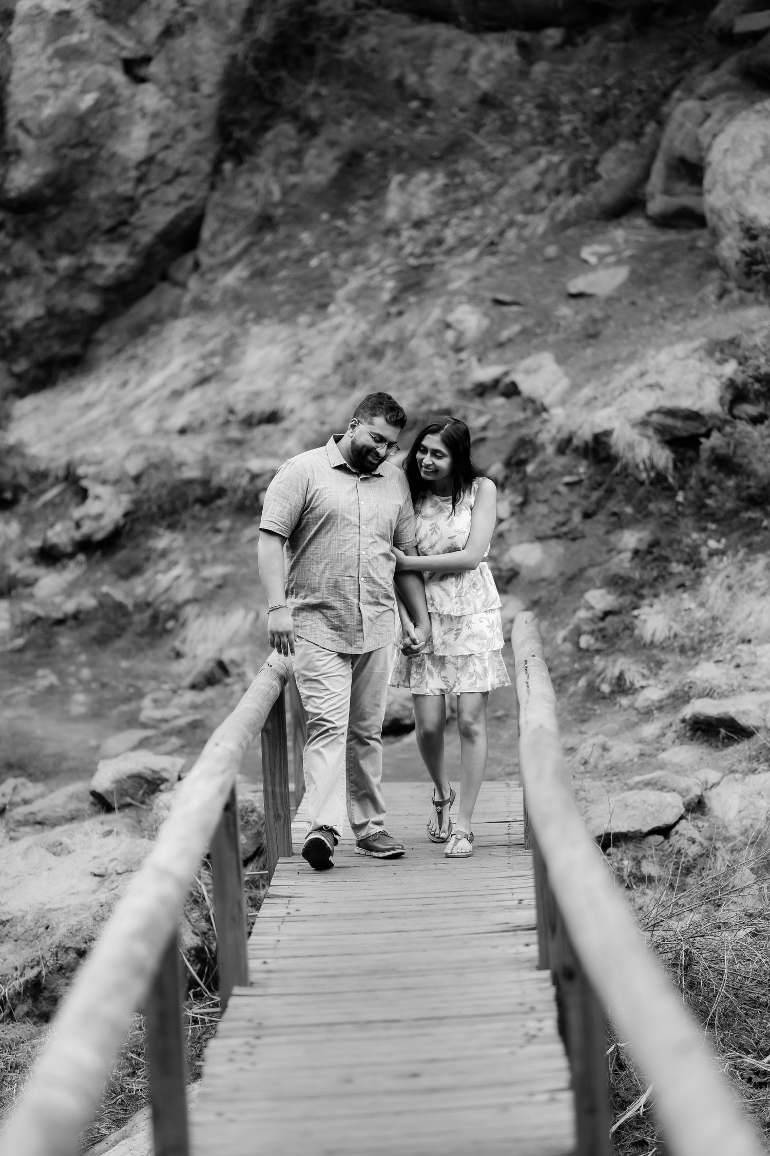 A couple walks together on a wooden pathway with rocky cliffs in the background.