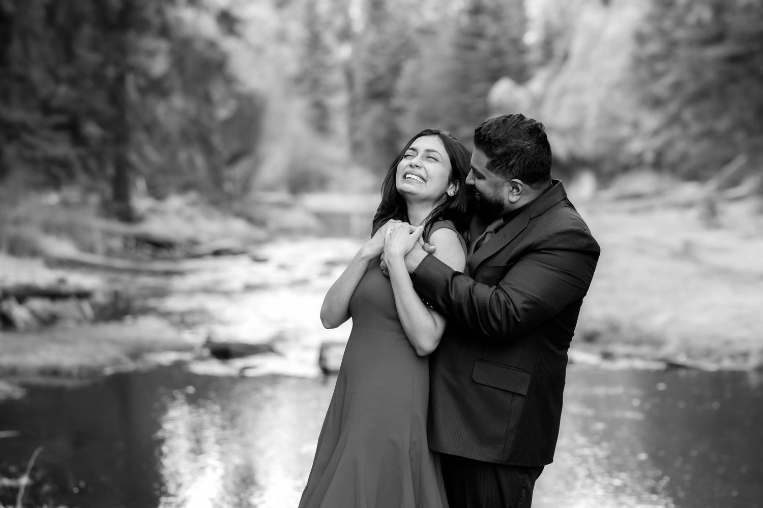A couple shares an intimate black and white moment by a river in a forest setting.