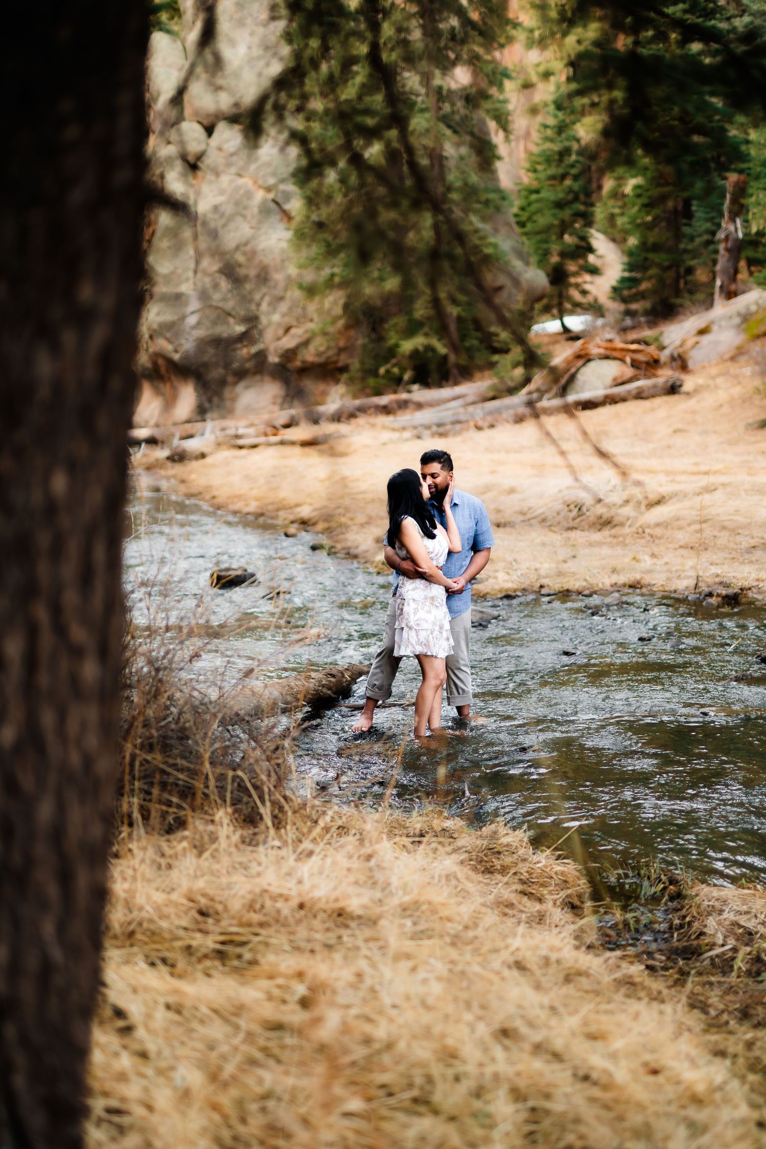A couple shares an intimate moment while standing in a stream with rocky cliffs and trees in background.