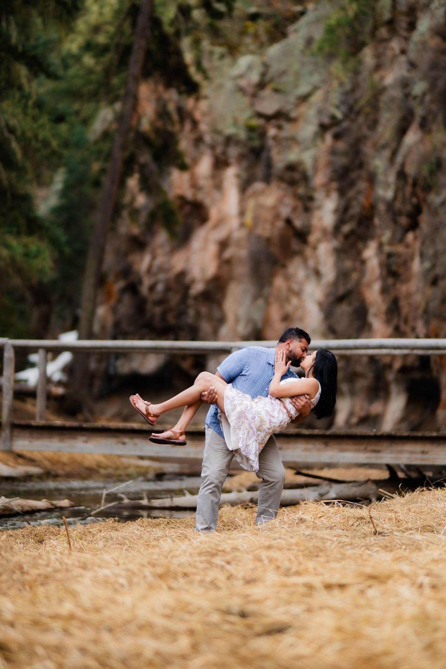 A playful couple shares a romantic dip pose on a wooden bridge in front of rocky cliffs.