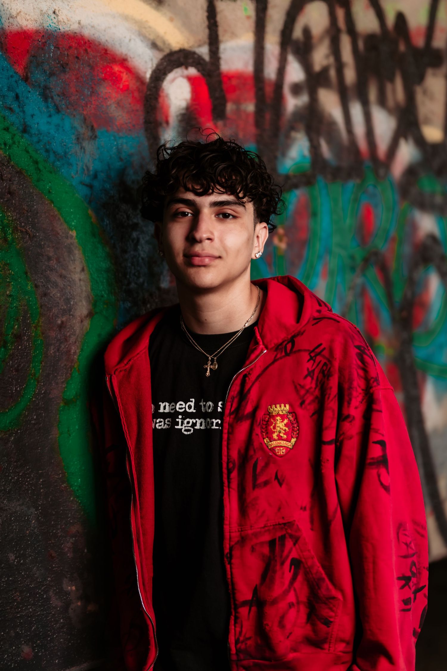 Portrait of someone in a red hoodie with emblem against graffiti wall in urban setting.