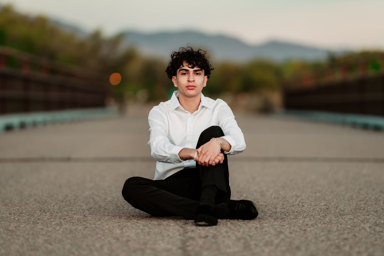 Person sitting cross-legged on concrete path with mountains and bokeh lights in background.