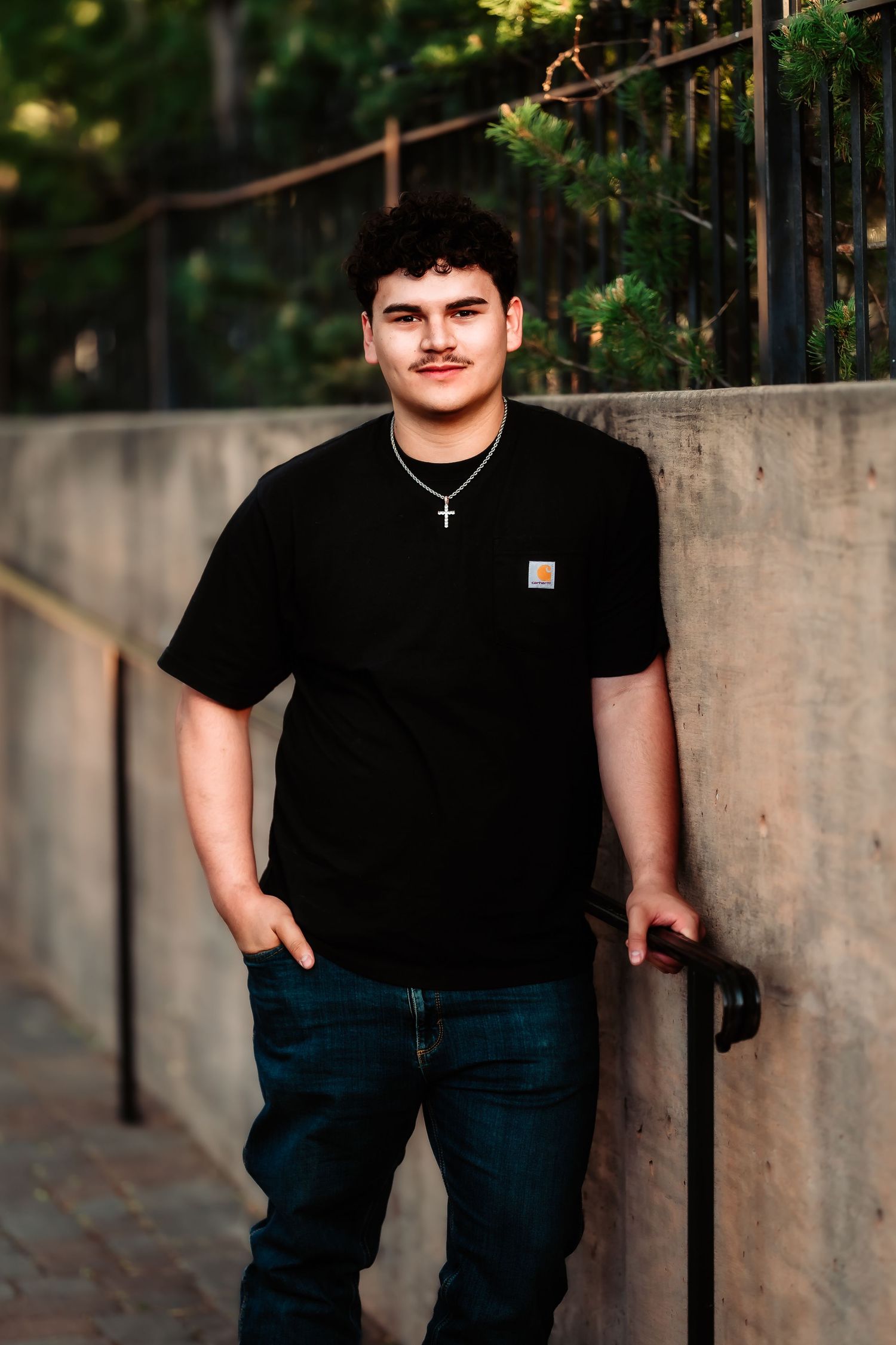 Portrait of a person in a black t-shirt with a cross necklace standing next to a concrete wall outdoors.