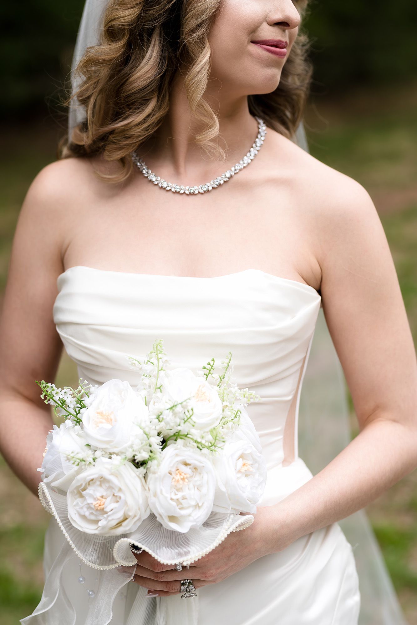Bride in strapless white gown holding elegant white rose bouquet, wearing diamond necklace and veil on her wedding day.