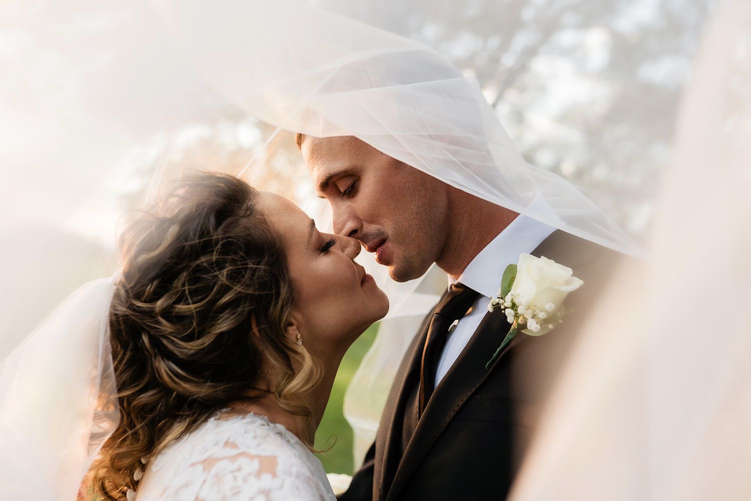 Romantic wedding couple share an intimate kiss outdoors with sunlight creating a dreamy atmosphere.