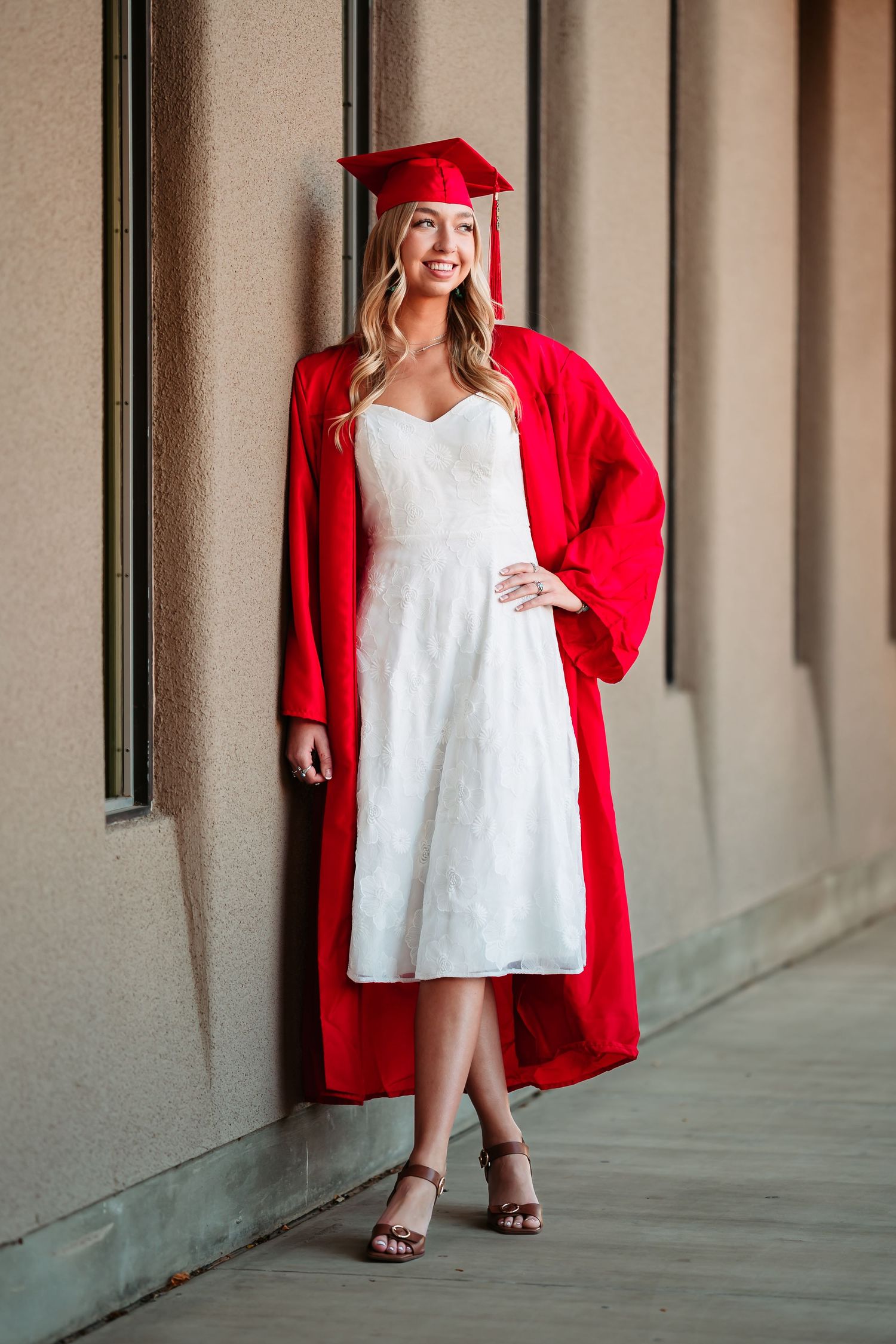 Graduate in red cap and gown poses in a white dress against a neutral hallway background for graduation photos.