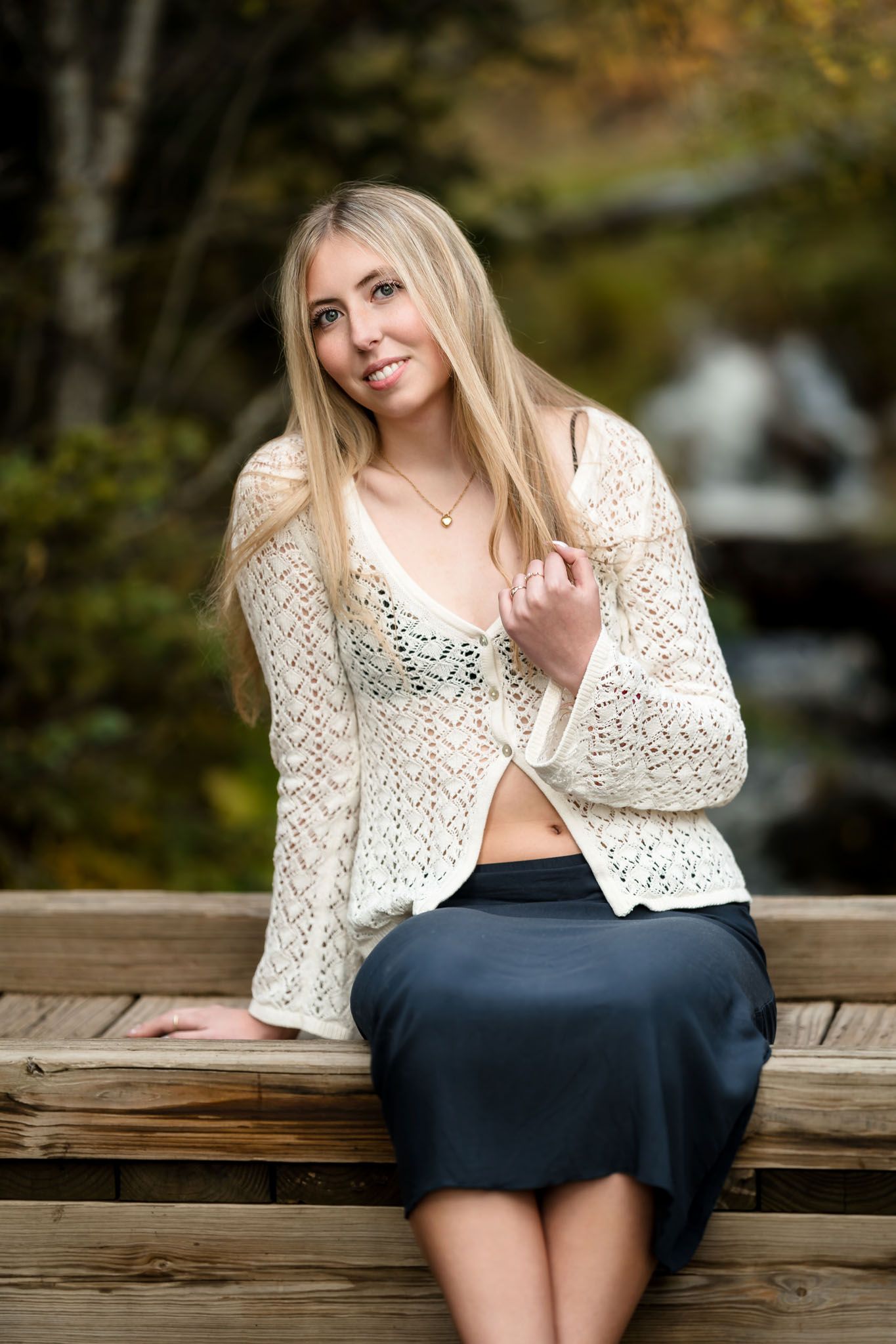 Blonde woman in white crochet cardigan sitting on wooden bench with waterfall backdrop in autumn.