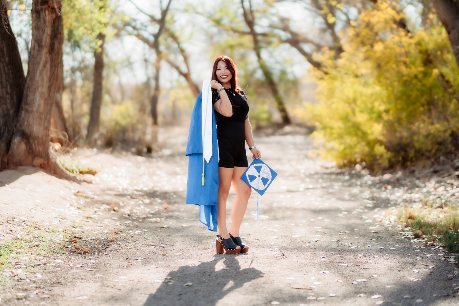 A person in a bright blue dress walks along a sunlit autumn path surrounded by golden trees and fallen leaves.