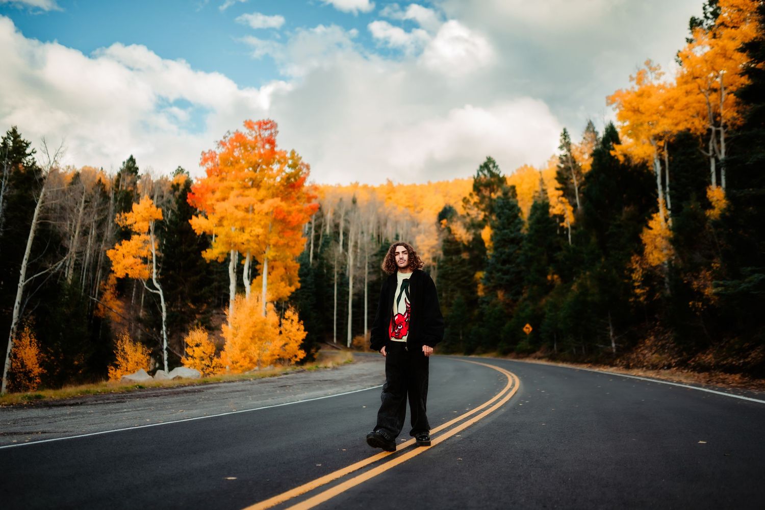 A winding mountain road curves through vibrant autumn trees with golden foliage against a cloudy sky.