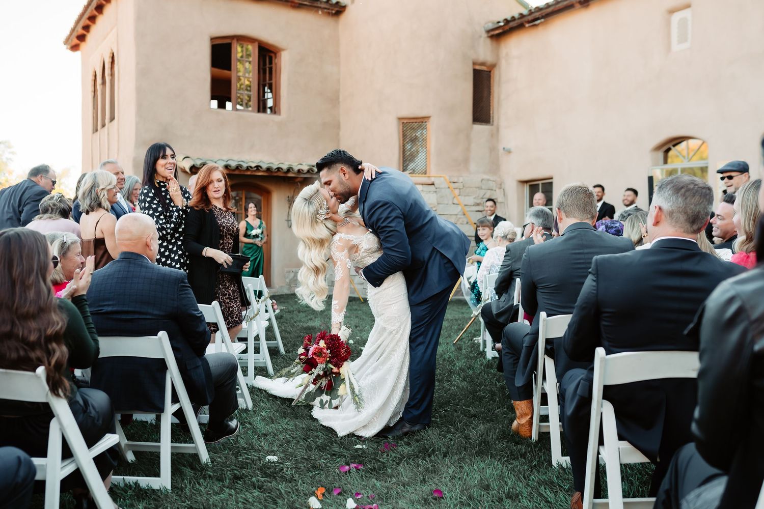 Wedding guests seated in white chairs at an outdoor ceremony venue with Spanish-style architecture in the background.
