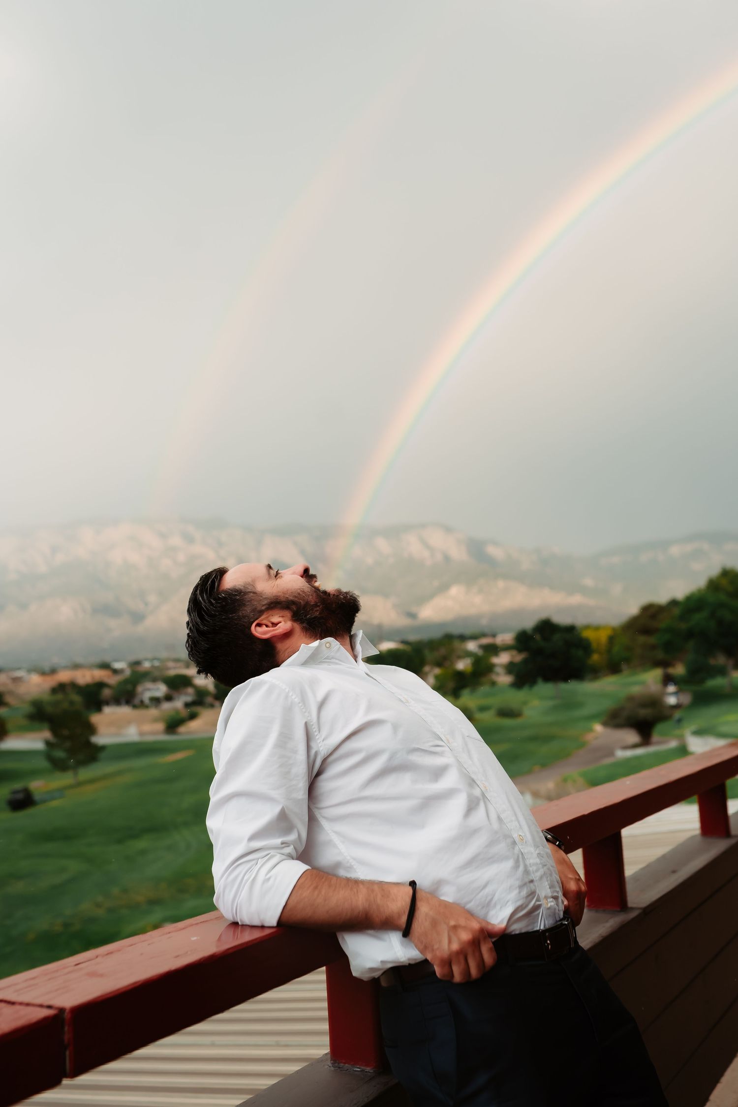 A person in a white shirt leans back on a wooden railing with mountains and a golf course visible in the background.