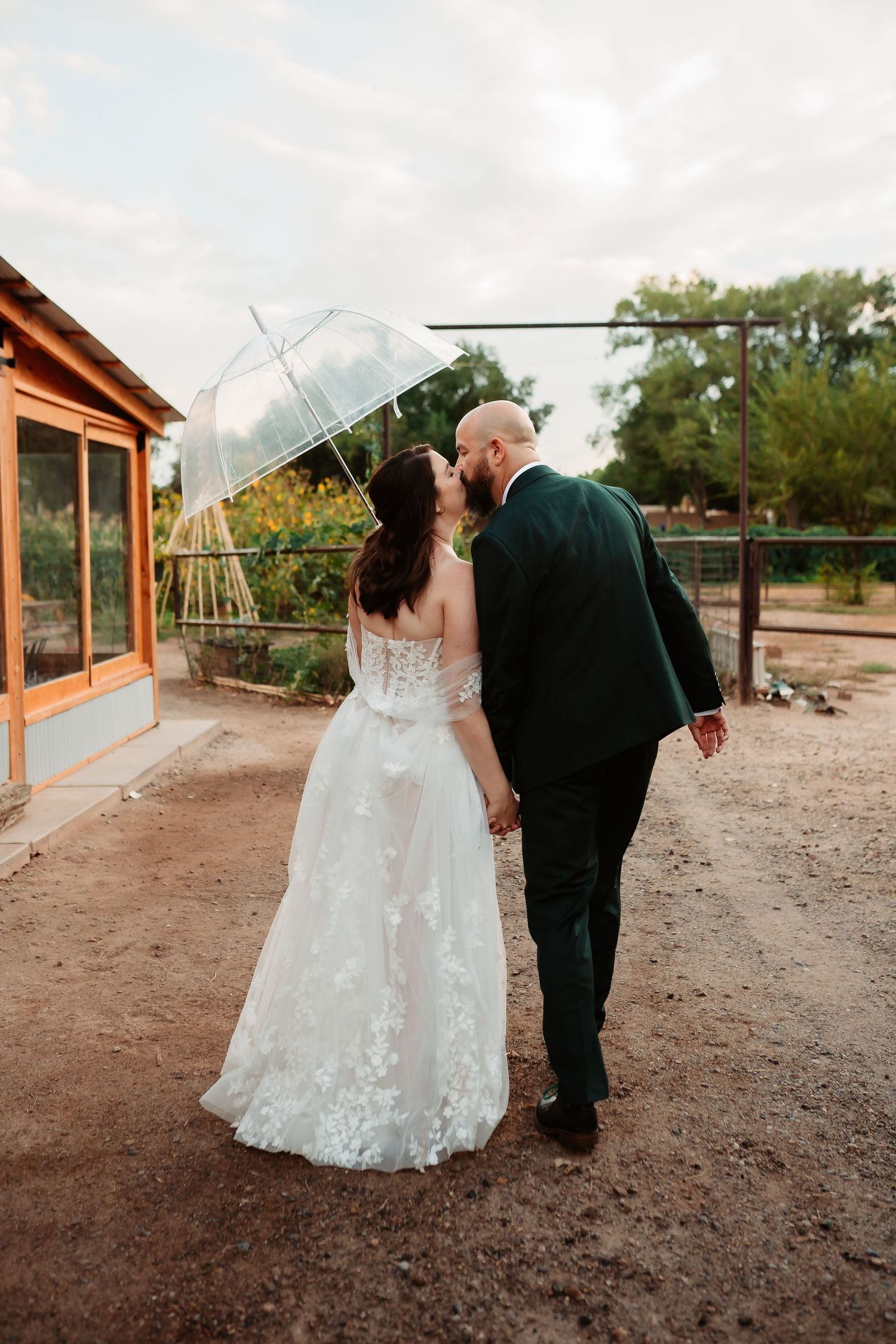 A couple in wedding attire walks together along a rustic outdoor venue with mountains in the background.